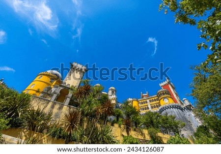 Wide angle exterior view of the Pena Palace decorated with mosaics and yellow and red painted walls over fortified walls covered in vegetation and palm trees under a sunny blue sky. Sintra. Portugal.