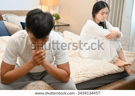 A sad, unhappy young Asian wife in pajamas is sitting separately with her husband on bed after their argument. relationship problems, misunderstanding, marriage conflict