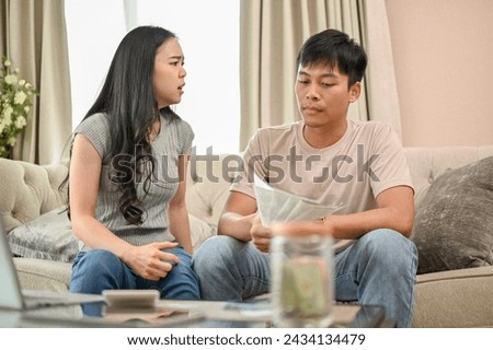 An unhappy and stressed Asian wife is arguing with her husband about unexpected expenses and high domestic bills on the sofa in the living room. spouses quarreling at home. family bankruptcy Royalty-Free Stock Photo #2434134479