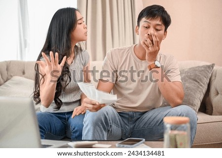 An unhappy and stressed Asian wife is arguing with her husband about unexpected expenses and high domestic bills on the sofa in the living room. spouses quarreling at home. family bankruptcy Royalty-Free Stock Photo #2434134481