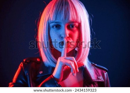 Finger by the lips, silence. Woman with white hair is in studio with neon colors.