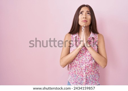 Young hispanic woman with long hair standing over pink background begging and praying with hands together with hope expression on face very emotional and worried. begging.  Royalty-Free Stock Photo #2434159857