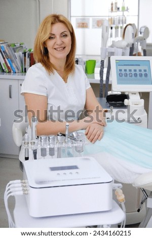 A friendly cosmetologist in a white medical suit smiles next to a professional mesotherapy machine with various treatment attachments in the clinic.