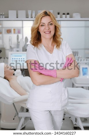 Confident female aesthetician in white uniform and pink gloves stands with arms crossed in a modern beauty clinic with a client behind her Royalty-Free Stock Photo #2434160227
