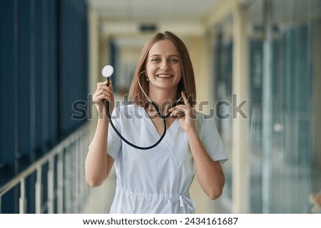 Using stethoscope. Female doctor in white coat is in the hall.