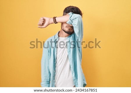 Young hispanic man with tattoos standing over yellow background covering eyes with arm, looking serious and sad. sightless, hiding and rejection concept  Royalty-Free Stock Photo #2434167851