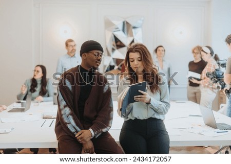 Young coworkers, a man and woman, deep in discussion holding a tablet with co-workers interacting in the background in a modern, creative office setting.