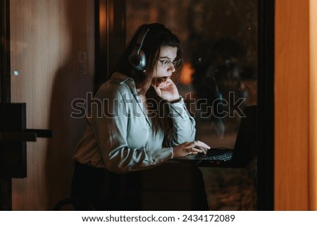 A dedicated young woman absorbed in her work on a computer with headphones, illuminated by her screen in a dark office setting. Royalty-Free Stock Photo #2434172089