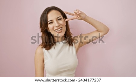 A cheerful young hispanic woman makes a peace sign against an isolated pink background, exuding confidence and beauty.