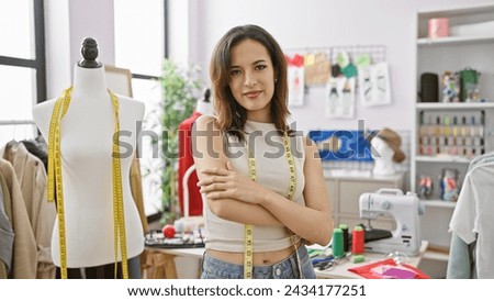 Hispanic woman with arms crossed stands in tailor shop with mannequin, sewing machine, and colorful threads.