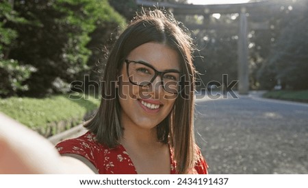 Cheerful, beautiful hispanic woman wearing glasses having fun making a confident selfie picture, smiling expression at meiji temple, tokyo's cultural shrine, capturing her happy moment.