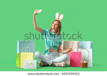 Young woman in bunny ears with shopping bags taking selfie on green background. Easter celebration