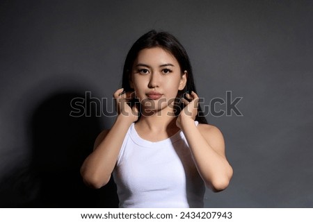 Self-confident young free business lady poses uninhibitedly in front camera, straightens hair with hand. Pretty Asian brunette looking straight at camera, hands raised hair, shadow on gray background
