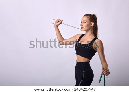 Young beautiful girl is a fitness trainer performing an exercise with a rope on a white background. A slender athletic athlete poses sideways with a skipping rope in her hand and looks away Royalty-Free Stock Photo #2434207947