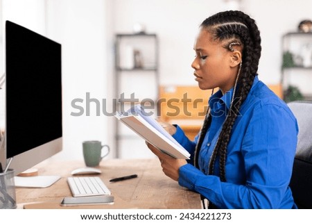 Studious teen female engrossed in reading textbook, preparing for an exam at her well-organized workspace in front of black computer monitor, side view Royalty-Free Stock Photo #2434221273