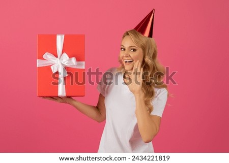 Attractive blonde lady wearing festive cap holds wrapped gift, whispering to camera great birthday offer, posing with excitement and joy on pink background. Bday and anniversary celebration