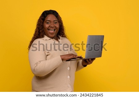 Cheerful overweight millennial black lady holding laptop computer looking at camera and exclaiming isolated on yellow studio background, checking online weigh loss course
