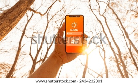 Hand holding smartphone with weather hot sunny day on screen and sunrise on midday at dry forest  background. High Temperature Weather show on Mobile Screen on Hot Sunny Day. concept global warming Royalty-Free Stock Photo #2434232301