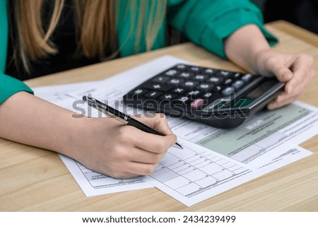 A woman sits at a desk, fills out a tax return and counts on a calculator Royalty-Free Stock Photo #2434239499