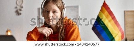 tired young teenage girl in wireless headphones looking at camera beside pride flag, banner Royalty-Free Stock Photo #2434240677