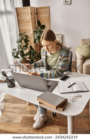 gen z girl holding book while using a laptop for e-learning at home, teen lifestyle concept Royalty-Free Stock Photo #2434242219