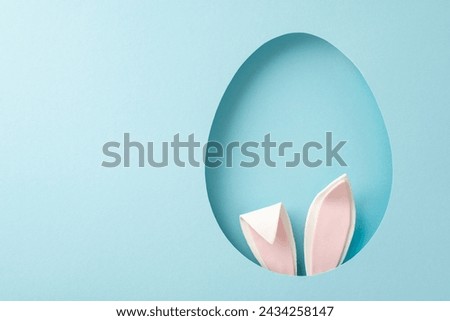 Playful Easter inspiration photo. Top view of humorous bunny ears peeking from an ovoid gap on a tranquil blue setting, with space for your text