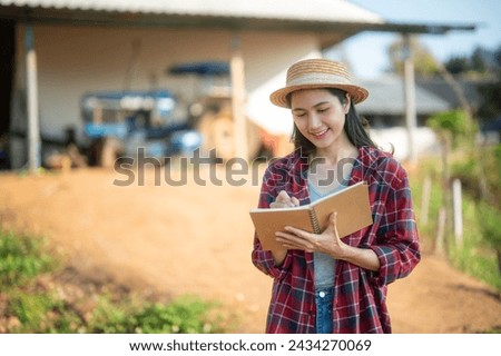 Proud female farmer stands watching and taking notes on the growth of her farm with a tractor behind her.