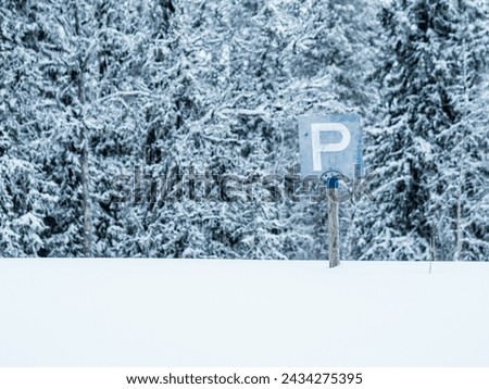 A parking sign stands prominently in the middle of a vast snowy field, surrounded by a winter landscape of white snow and bare trees.