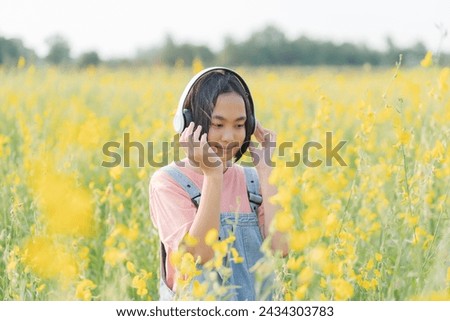 A young Asian girl happily listens to music using headphones in a field of yellow flowers.
