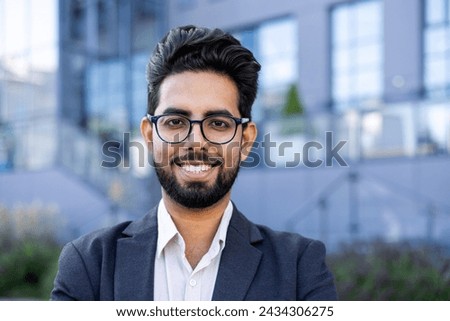 A young Indian businessman in a sharp suit stands confidently outdoors with an urban landscape in the background. Ideal for corporate diversity themes. Royalty-Free Stock Photo #2434306275