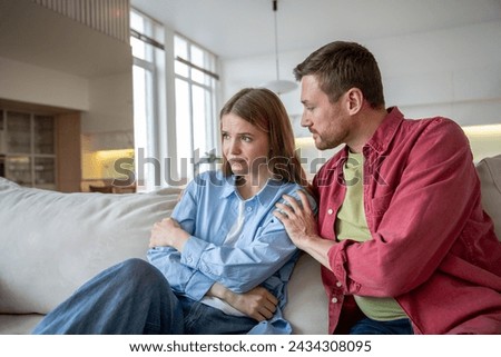 Offended woman sits with arms crossed and frowning, turning back man. Nearby, apologetic husband puts arm around shoulders trying to look into eyes. Young couple is ready to make peace after quarrel. Royalty-Free Stock Photo #2434308095