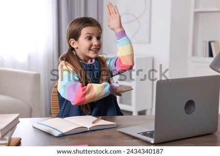 E-learning. Emotional girl raising her hand to answer during online lesson at table indoors