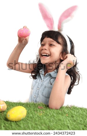 Cute little girl with pink bunny ears painting eggs with brush on green grass meadow. Joyful kid celebrating Easter day on white background. Happy child having fun on Easter holiday celebration.