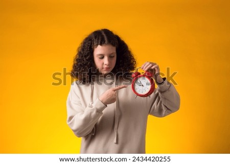 Portrait of smiling girl holding an alarm clock in hand and pointing finger at it. The concept of education, school, deadlines, time to study