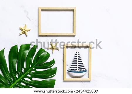 Summer vacation concept. Artistic composition with big tropical monstera leaf, decorative star fish and  wooden picture frame with a boat on white marble background. Copy space for your design.
