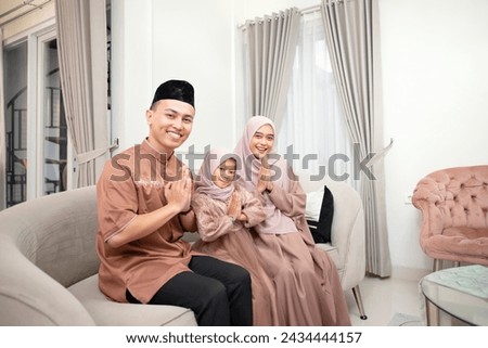 Young Indonesian muslim family, with greetings gesture, welcoming guests celebrating Eid in traditional outfit at home. Muslim, ramadan and islamic concept. Royalty-Free Stock Photo #2434444157