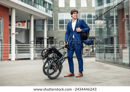 Male businessman wearing a blue suit poses confidently with his folded electric folding bike in the bustling financial district of the city, carrying a backpack and wearing a bike helmet for safety