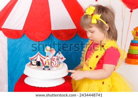 A little girl in a colorful outfit is touching a birthday cake. Circus theme, white, blue, red, yellow colors. Horizontal picture. 