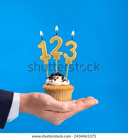 Hand holding a cupcake with the number 123 candle - Birthday on blue background