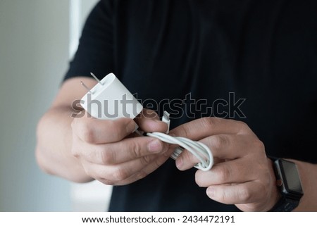 close up of the hands of a man holding a white cell phone charger and carrying a watch on his left arm. he wears a black t-shirt