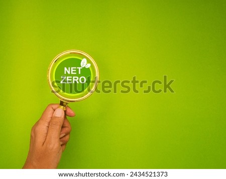 Hand holding a magnifying glass on a green background with text Net Zero. Top view. Net zero gas emissions target.
