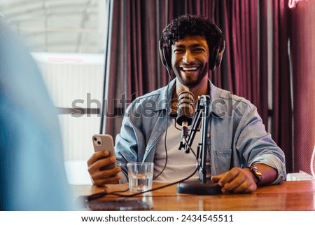 Happy podcaster in a casual denim jacket smiling while recording a podcast. He sits at a studio table with a microphone, wearing headphones and holding a smartphone, displaying a lively personality.