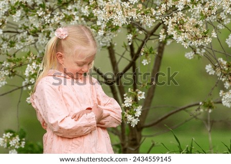 Cute blonde girl in blooming garden hugging herself. Little princess in pink dress stands near flowering tree.