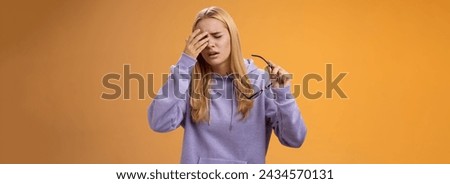 Tired dizzy cute blond girl taking off glasses touching forehead feel sick unwell suffering headache painful migraine standing bothered uncomfortable orange background in hoodie.