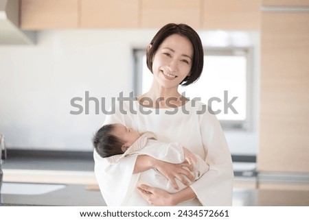 A Japanese mother smiles for the camera as she holds her infant in the living room. Royalty-Free Stock Photo #2434572661