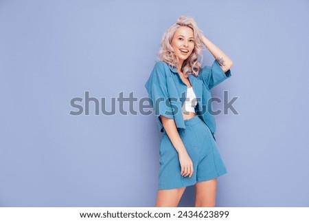 Young beautiful smiling female in trendy summer blue costume clothes. Carefree blond woman posing near violet wall in studio in jacket and shorts. Positive model having fun. Cheerful and happy