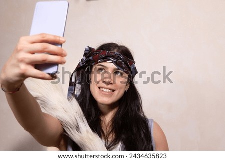 A vivacious young woman adorned with a stylish headscarf smiles as she captures a selfie using her smartphone, exuding a sense of joy and contemporary fashion Royalty-Free Stock Photo #2434635823