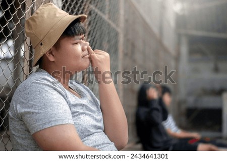 Asian teenboy in white t-shirt sits with picking mucus against a metal fence panel in a juvenile detention facility, awaiting further release, freedom and detention of people concept. Royalty-Free Stock Photo #2434641301
