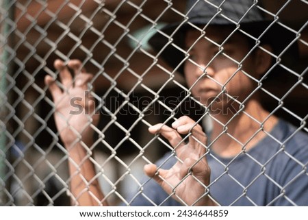 A young boy wearing a gray hat and t-shirt stands with his hands on a metal fence in a detention center, looking sad and wanting his freedom. Royalty-Free Stock Photo #2434644859