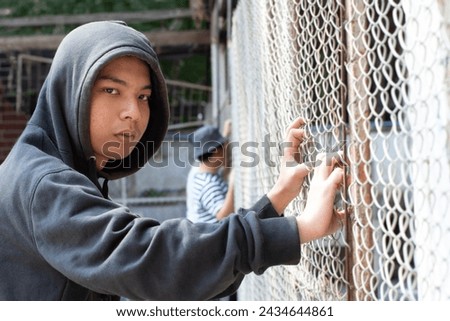 A young boy wearing a gray hoody stands with his hands on a metal fence in a detention center with his friends, looking sad and wanting his freedom. Royalty-Free Stock Photo #2434644861